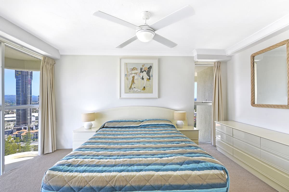A hotel room with a bed, a ceiling fan, a framed artwork, a dresser, and a view of the city.