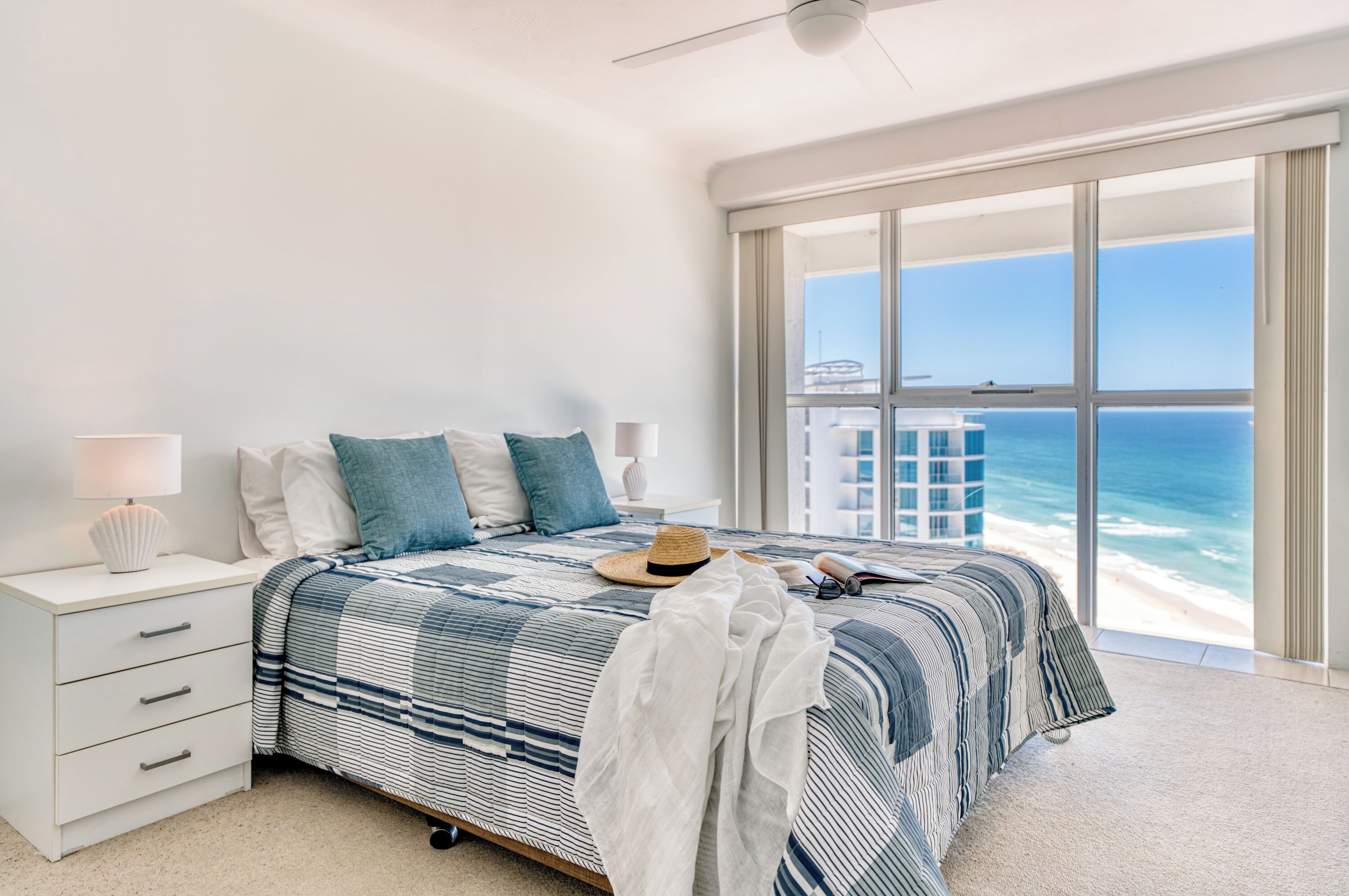 A bedroom with a large window overlooking the ocean, a bed with striped bedding, a nightstand with a lamp, and a suitcase on the bed.