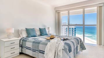 A bedroom with a large window overlooking the ocean, a bed with striped bedding, a nightstand with a lamp, and a suitcase on the bed.