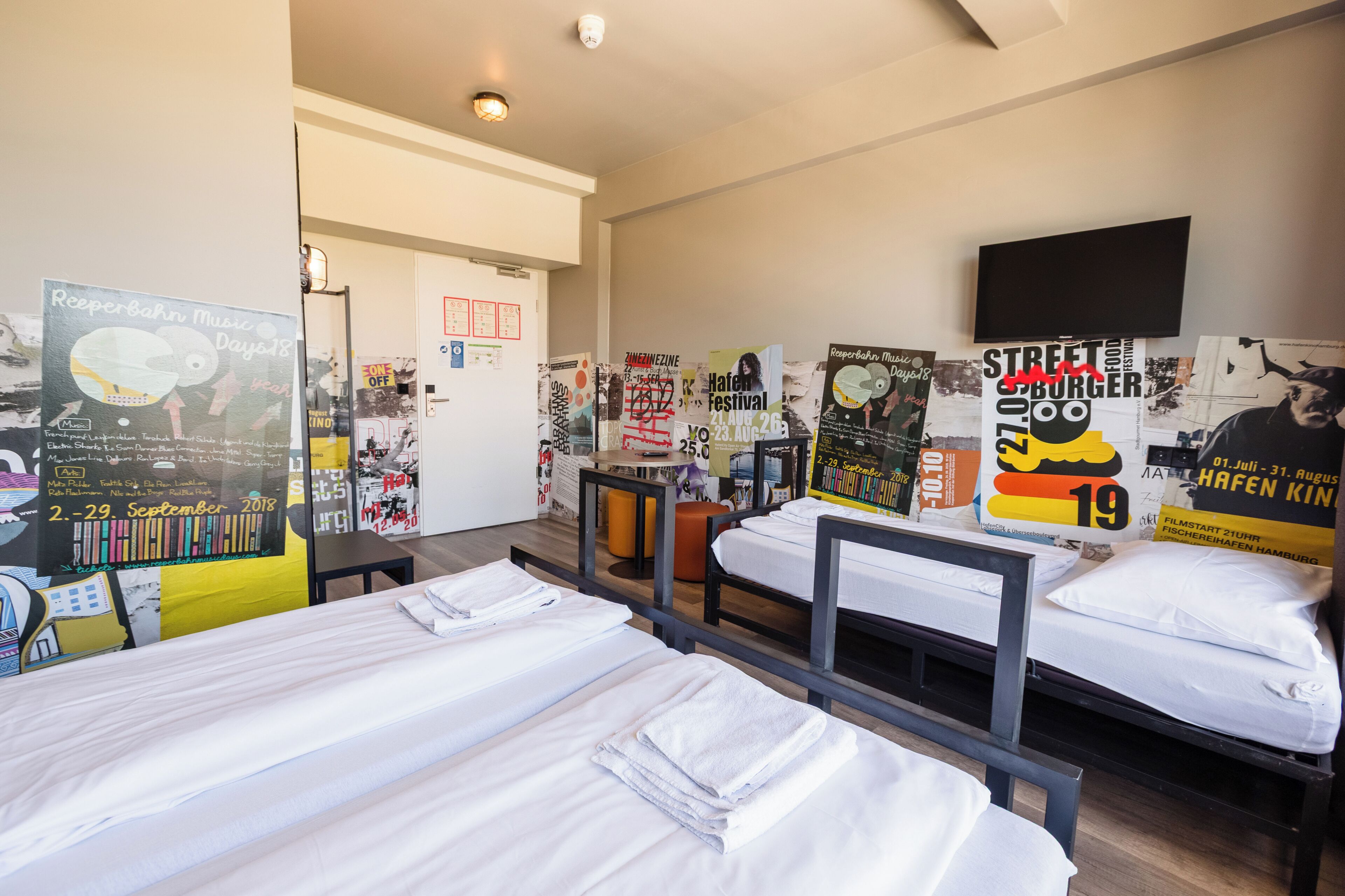 Une chambre d’hôtel avec trois lits, un téléviseur à écran plat et divers affiches aux murs.