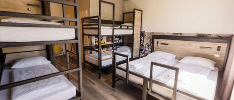 A dormitory-style room with bunk beds, a wooden floor, and a small shelf with books.