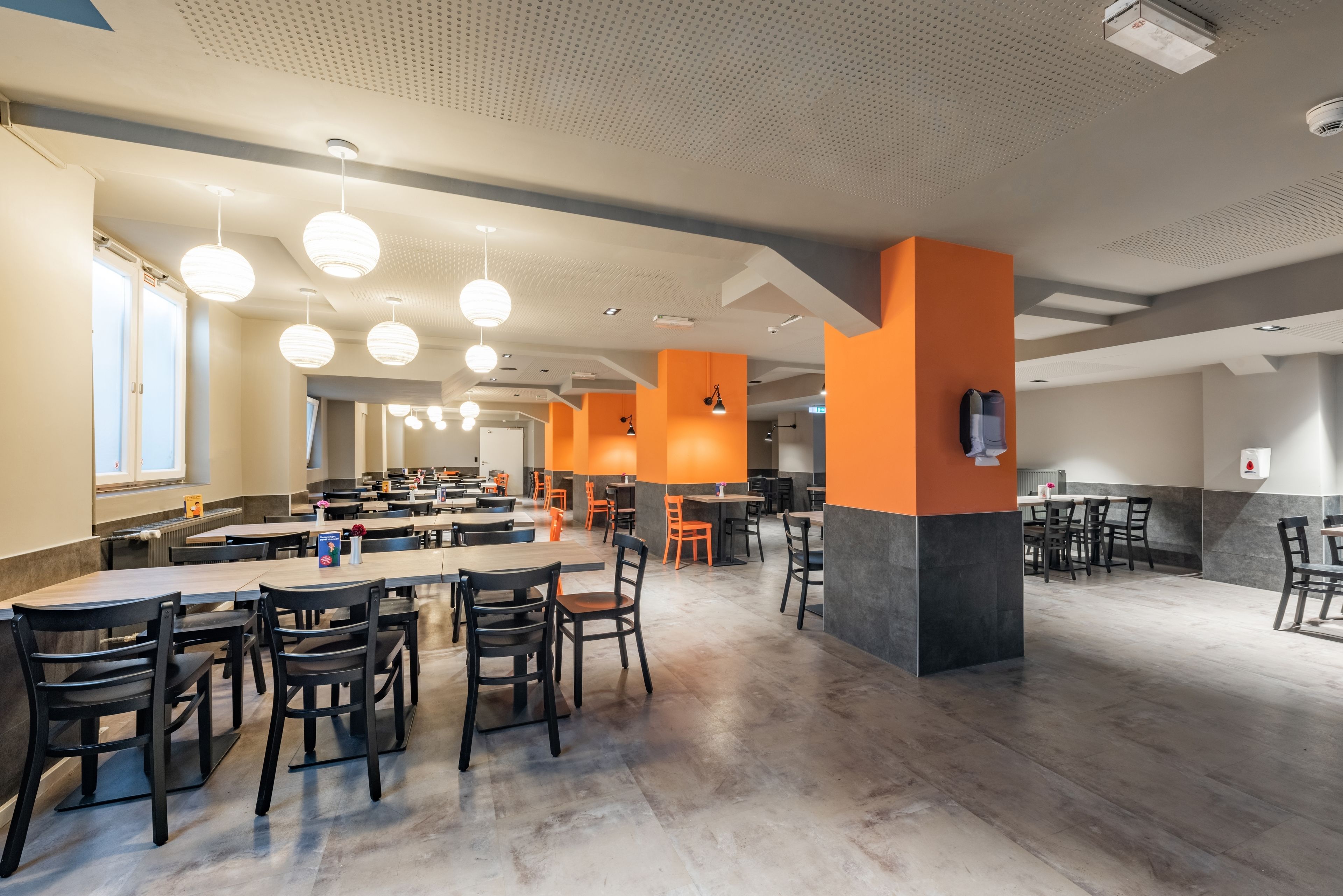 A spacious cafeteria with multiple tables and chairs, featuring a modern design with orange accents and a high ceiling with pendant lighting.
