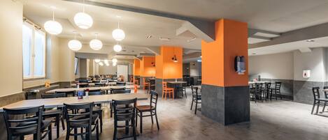 Una amplia cafetería con varias mesas y sillas, con un diseño moderno y detalles en naranja, y un techo alto con iluminación colgante.