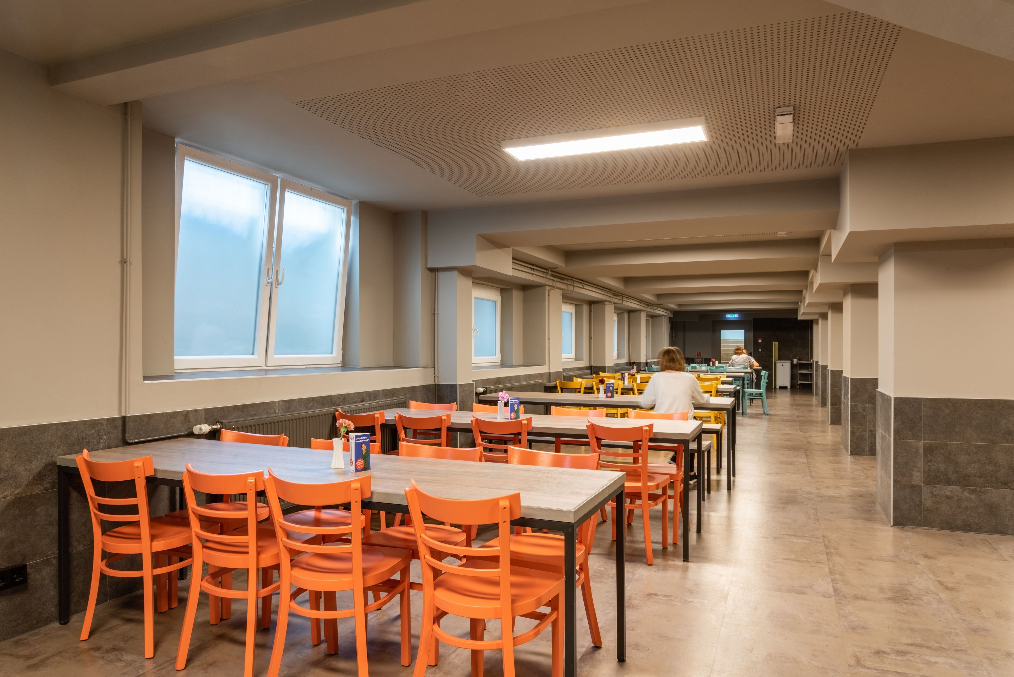 Una cafetería amplia con mesas largas y sillas naranjas, ventanas grandes y piso de baldosas.