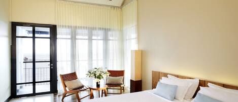 Habitación de hotel con una cama grande, dos sillas, una mesita con una planta y una puerta al balcón.