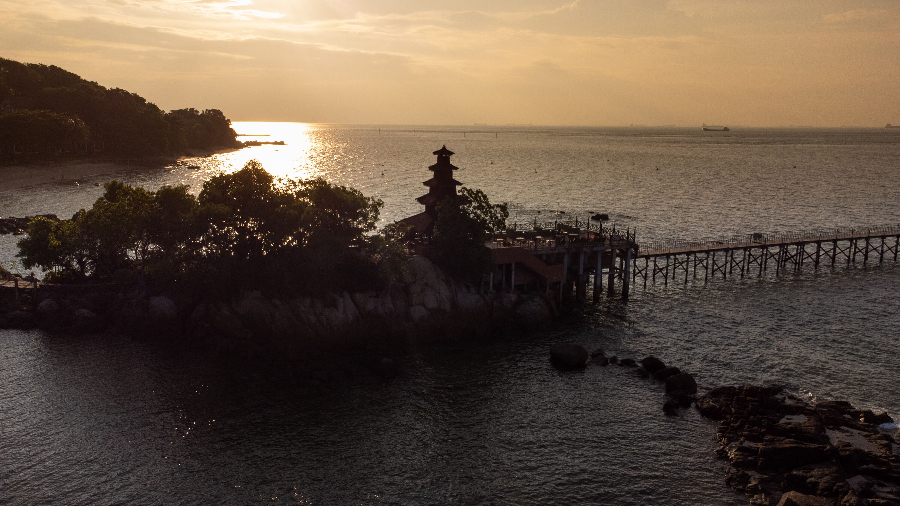 A coastal scene with a pier extending into the sea, a small island with a traditional structure, and a sunset in the background.