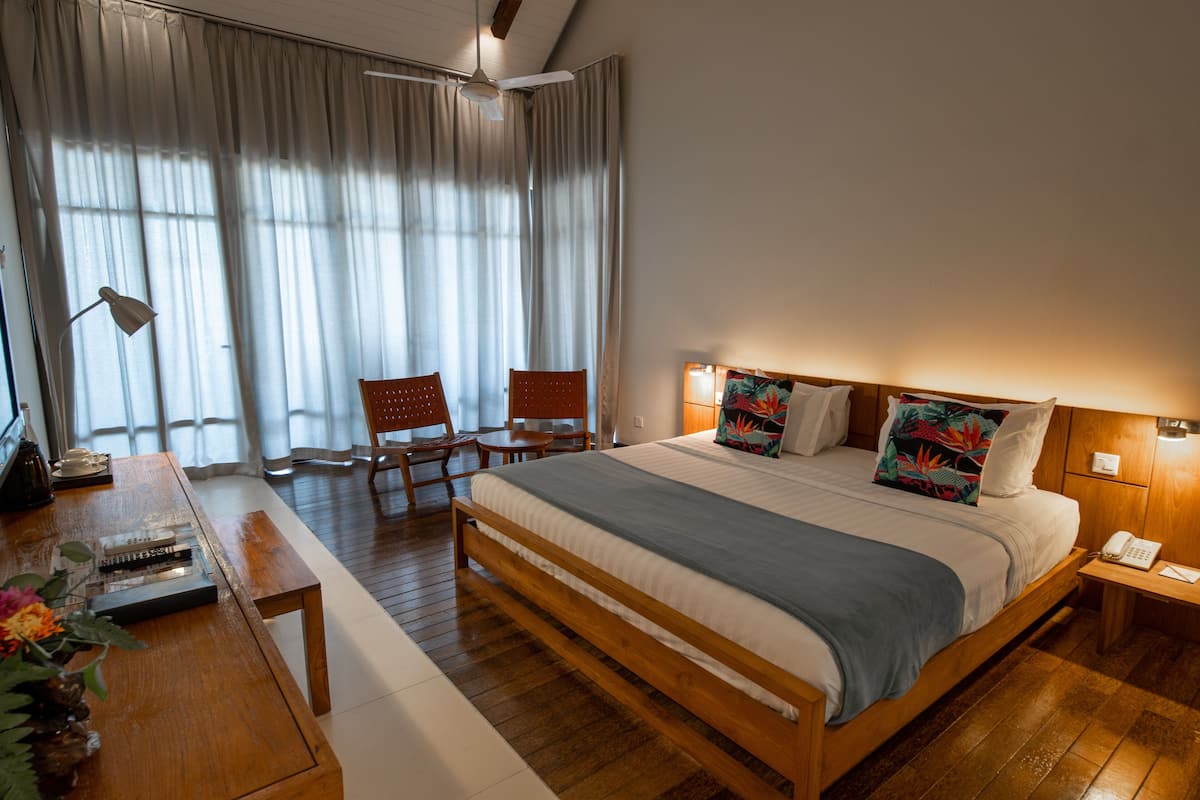 Ein Hotelzimmer mit einem hölzernen Bett, einem Schreibtisch mit Lampe, einem Stuhl und einem großen Fenster mit Vorhängen.