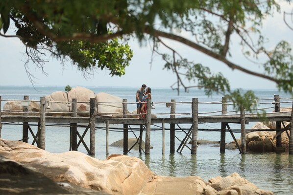 A wooden pier extending over calm water, with large rocks in the foreground and a couple standing on it. - Turi Beach Resort (Batam)