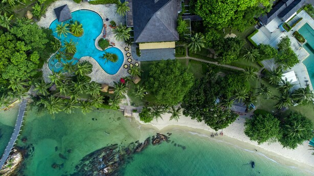 Vista aérea de un resort con una gran piscina, palmeras y una zona de playa.