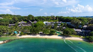 Vista aérea de un complejo turístico costero con villas frente a la playa, una playa de arena y aguas turquesas cristalinas.