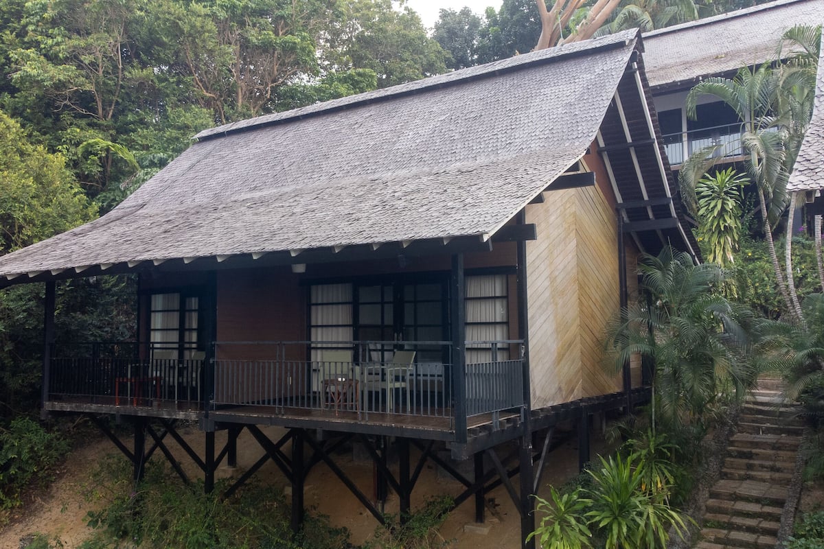 Ein traditionelles Holzhaus mit einem strohgedeckten Dach, das auf Stelzen steht, umgeben von dichtem Grün.