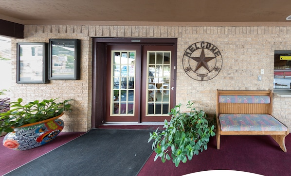 A hotel entrance with a brick wall, a welcome sign, a bench, and a potted plant. - Americas Best Value Inn The Legends Inn (Junction)
