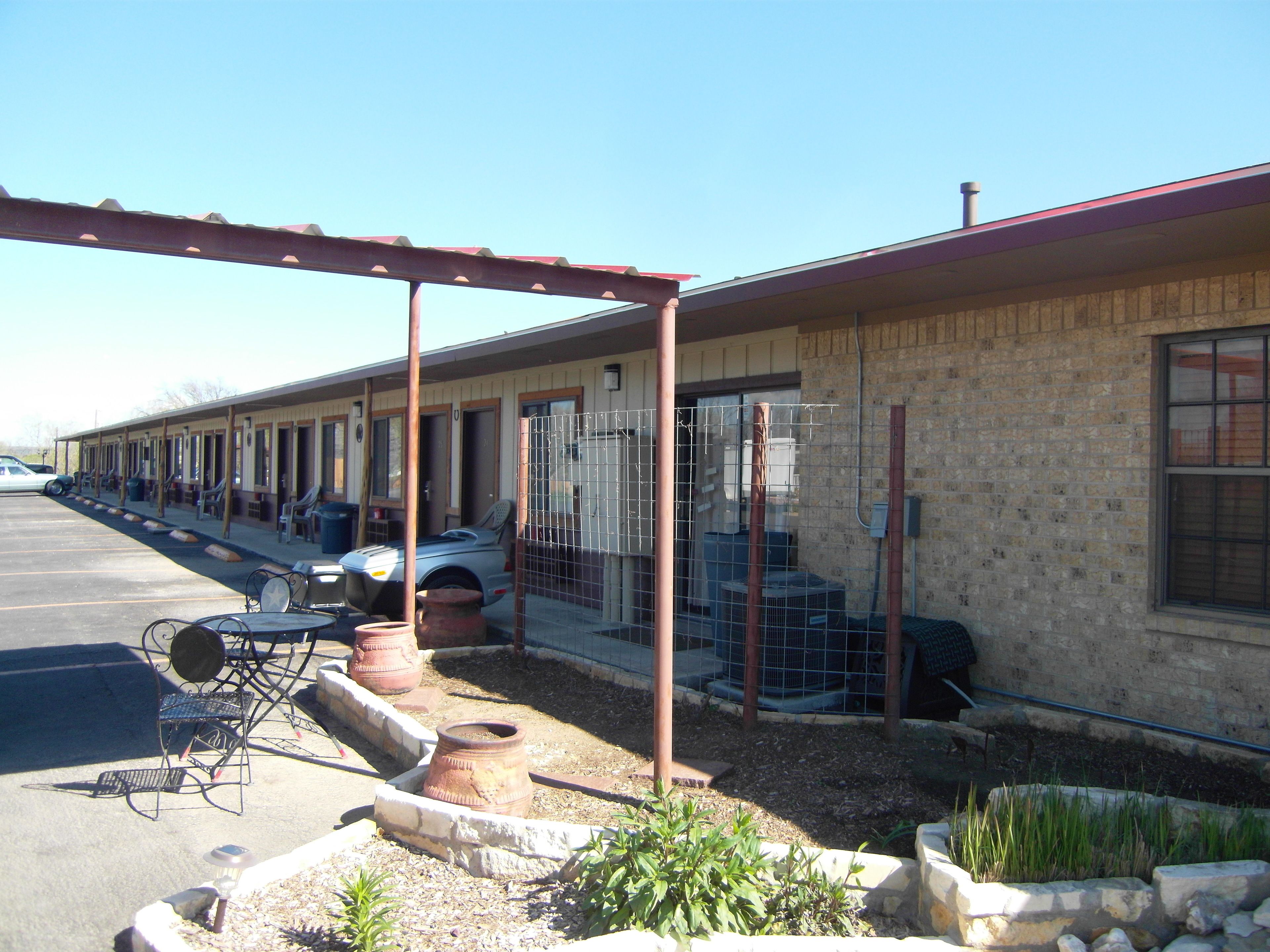 A motel with a row of rooms, a covered parking area, and a small garden with plants.