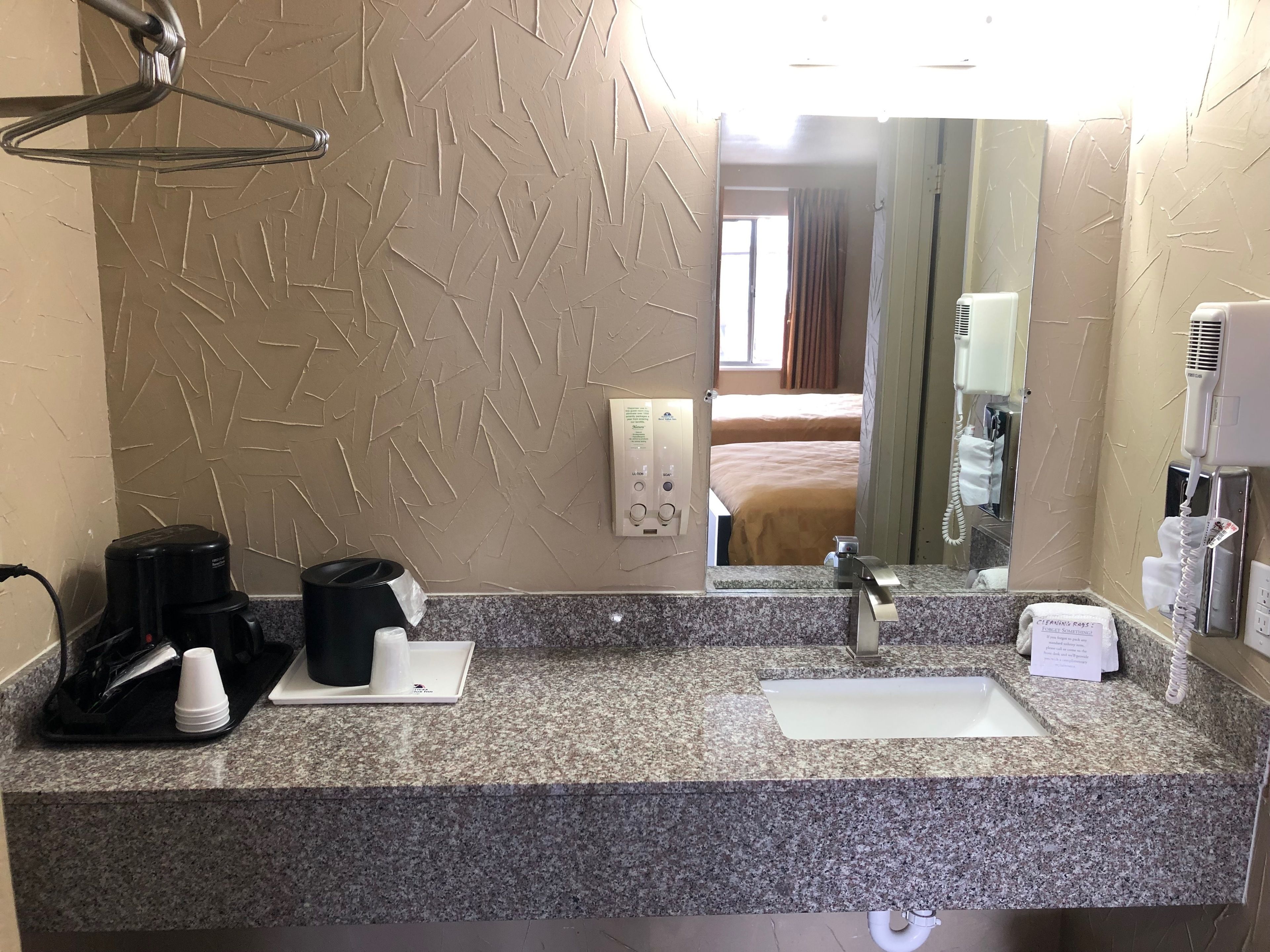Ett hotellbadrum med en bänkskiva i granit, ett handfat, en spegel, en handduksdispenser, en hårtork och en kaffebryggare.