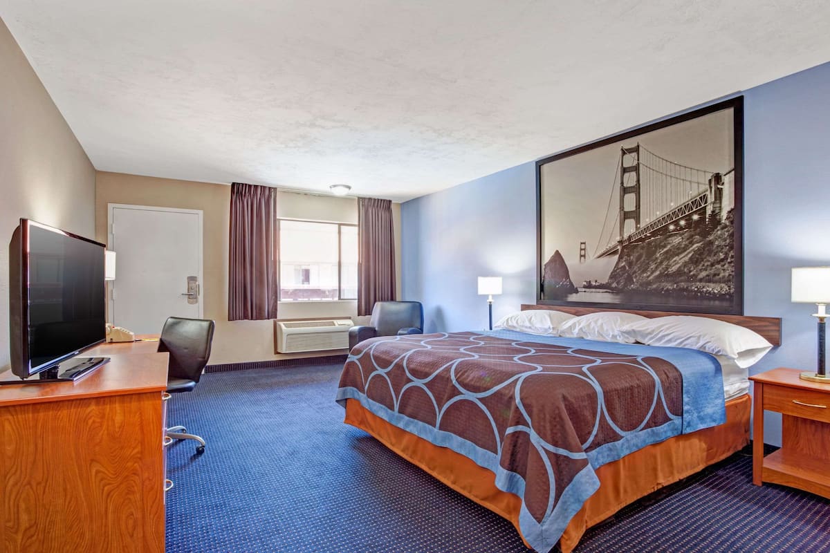 A hotel room with a bed, a desk with a TV, a chair, a desk lamp, a large window with curtains, and a wall-mounted air conditioning unit.
