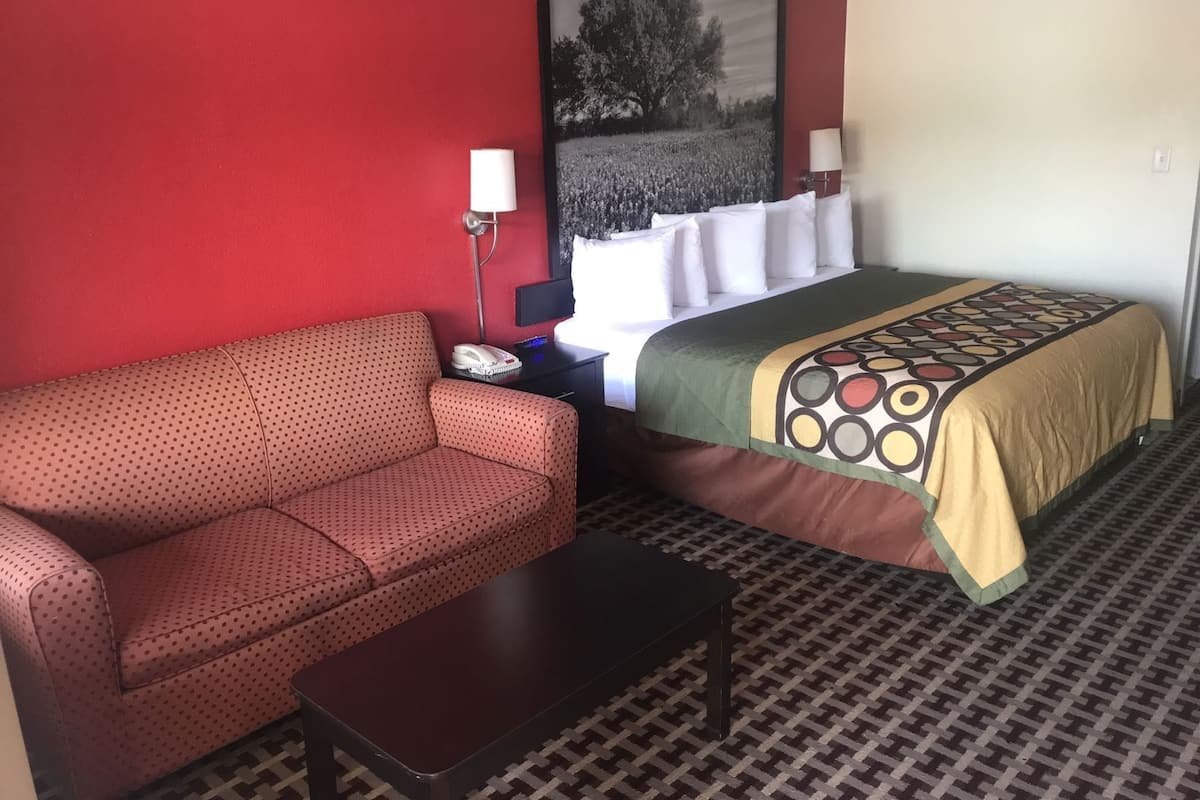 A hotel room with a red wall, a bed with a patterned bedspread, a sofa, a small table, and a nightstand with a lamp.