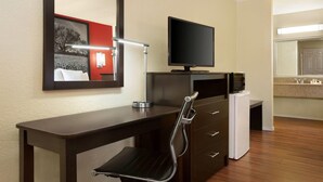 A hotel room with a dark wooden desk, a modern chair, a flat-screen TV, a small refrigerator, and a mirror on the wall.
