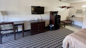 A hotel room with a bed, a desk, a dresser, a television, a microwave, a trash bin, and a bathroom with a sink and mirror.