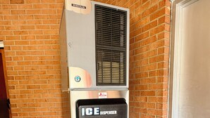 A vending machine with a 'HOSHIZAKI' label and an 'ICE DISPENSER' below it, placed against a brick wall with a tiled floor.