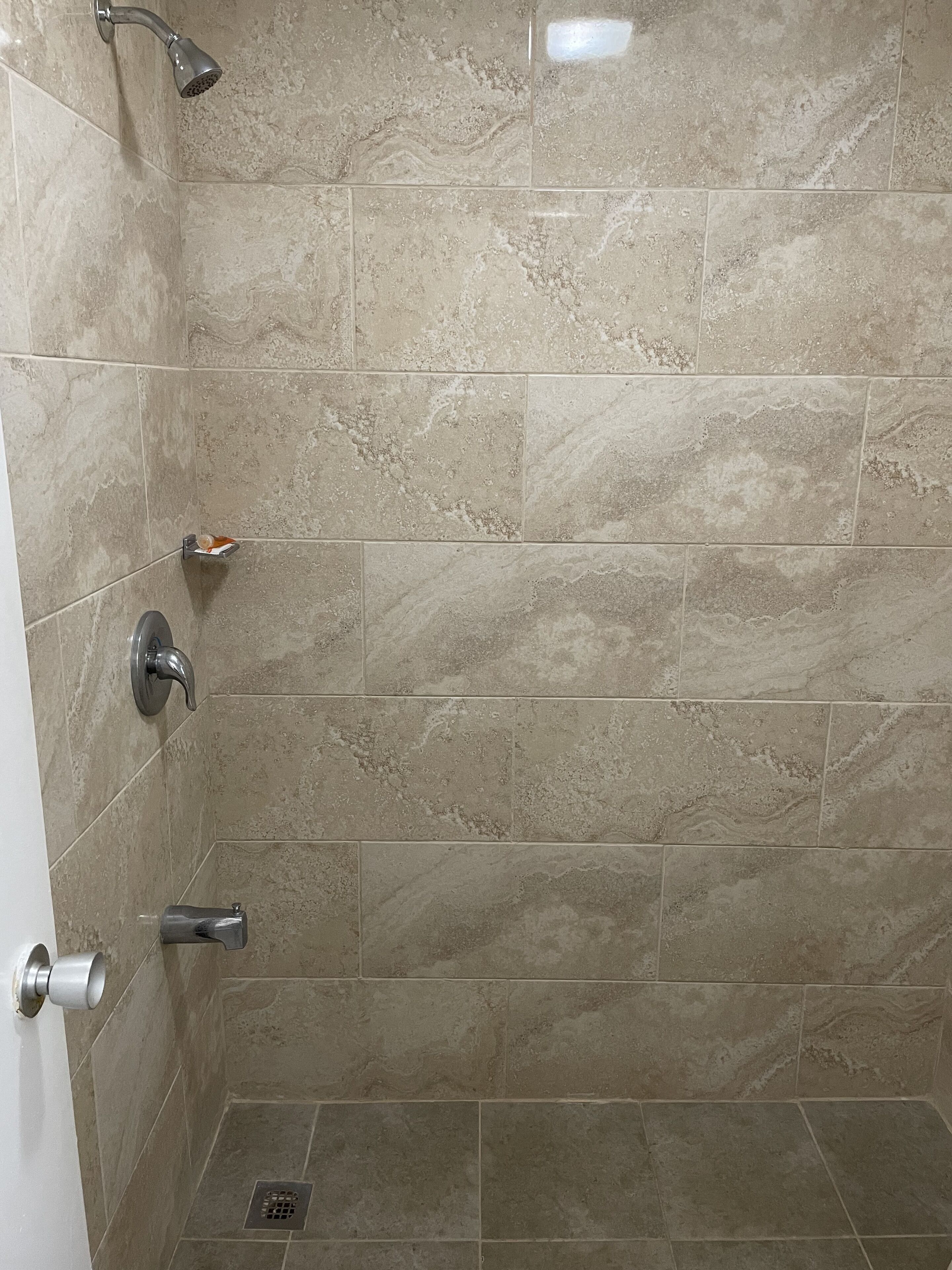 Un espace douche avec des carreaux de carrelage beige, une douchette à main, un robinet mural et un drain au sol.