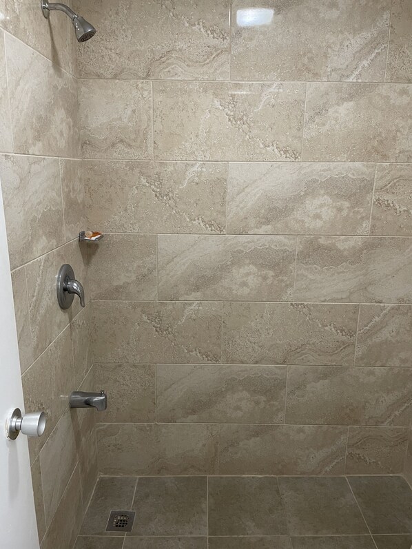 A shower area with beige tiles, a handheld showerhead, a wall-mounted faucet, and a floor drain. - Washington Burgess Inn (West Point)