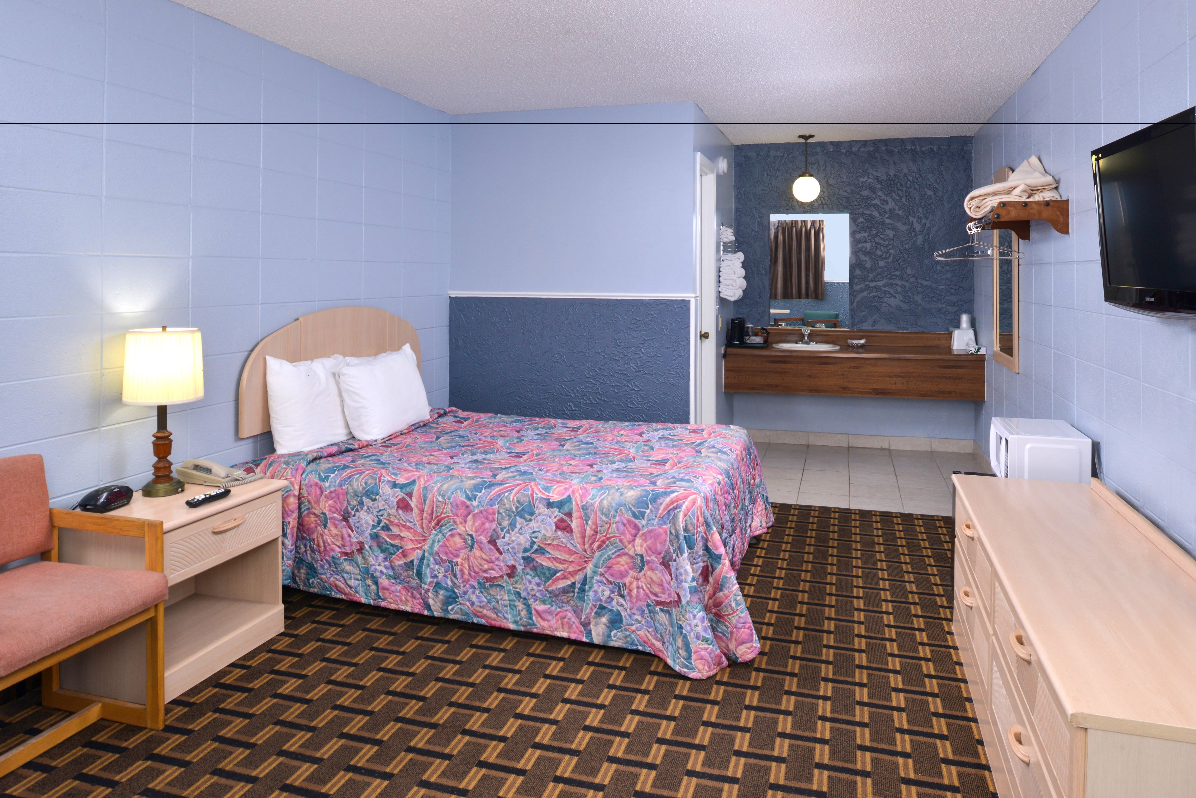Ein Hotelzimmer mit Bett, Schreibtisch mit Lampe, Stuhl, Kommode, Fernseher und einem Badezimmer mit Waschbecken und Spiegel sowie einem gemusterten Teppich.