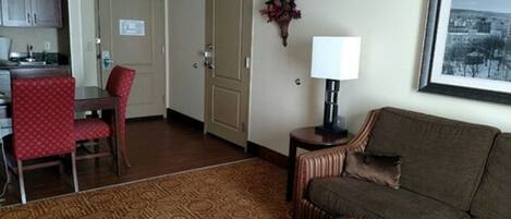 A hotel room with a dining area, a sofa, a lamp, and a view of the city.