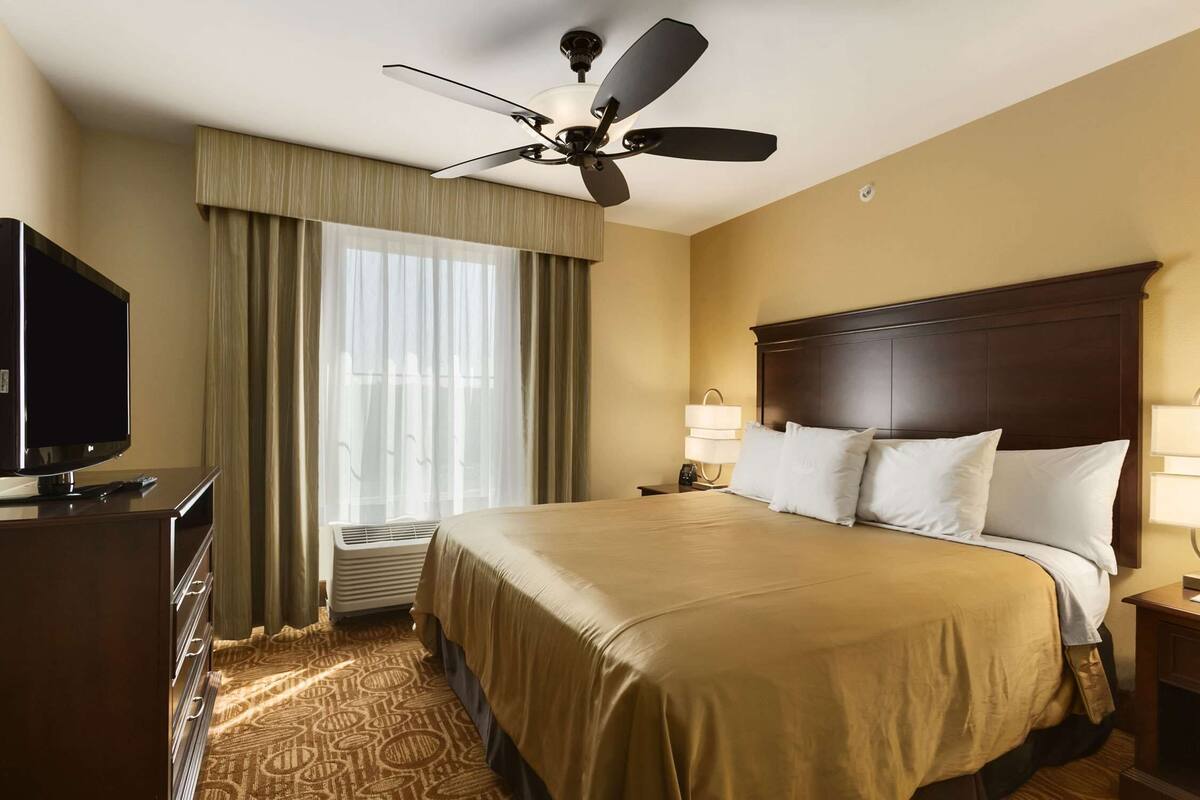 Habitación de hotel con una cama grande, ventilador de techo, televisor sobre un aparador y una ventana con cortinas.
