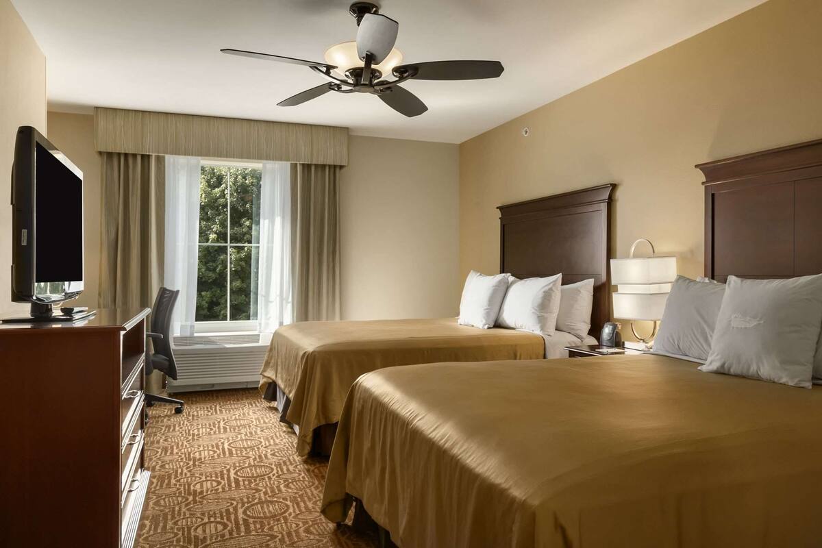 Habitación de hotel con dos camas, ventilador de techo, televisión, escritorio y ventana con cortinas.