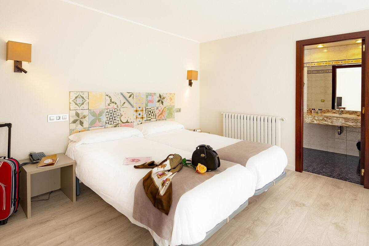 Une chambre d’hôtel avec un grand lit, une table de chevet, une valise rouge et une salle de bain visible à travers une porte ouverte.