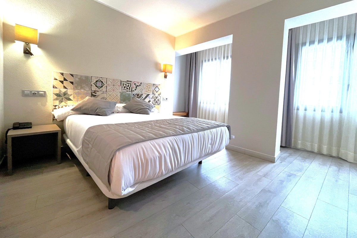 Une chambre d’hôtel avec un lit, des tables de chevet, une lampe fixée au mur et une fenêtre avec des rideaux.