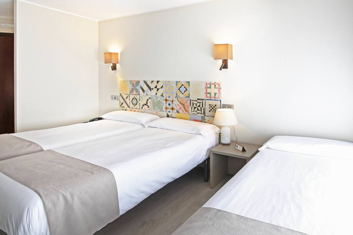 Une chambre d’hôtel avec deux lits, une table de chevet, une lampe et un mur décoré d’une mosaïque colorée.