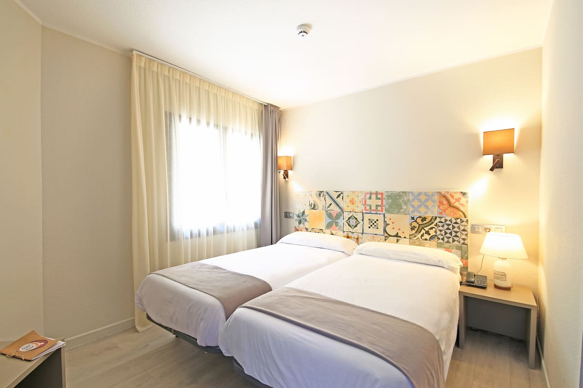 Une chambre d’hôtel avec deux lits, une table de chevet avec une lampe, une fenêtre avec des rideaux et un mur orné d’un motif de carreaux colorés.