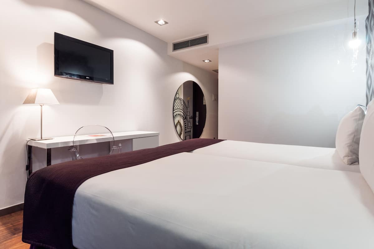 Moderni hotellihuone, jossa on suuri sänky, työpöytä lampun kanssa, seinälle asennettu taulutelevisio ja koristeellinen peili.