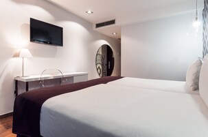 Quarto de hotel moderno com uma cama grande, uma escrivaninha com luminária, uma TV de tela plana na parede e um espelho decorativo.