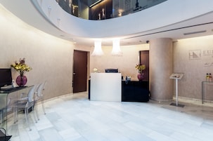 Un área de recepción de hotel moderna con techo curvo, mostrador de recepción, sillas transparentes y una pantalla digital.