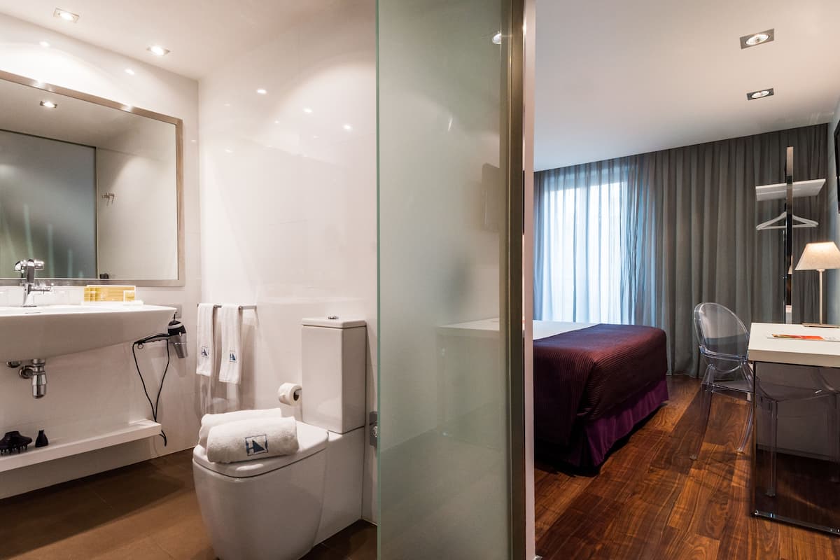 Moderni hotellihuone, jossa on lasiseinä, joka erottaa kylpyhuoneen ja makuuhuoneen. Kylpyhuoneessa on valkoinen pesuallas, wc ja suihkutila. Makuuhuoneessa on sänky, työpöytä tuolilla ja ikkuna, jossa on verhot.