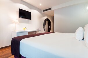 A modern hotel room with a large bed, a desk with a lamp, and a flat-screen TV mounted on the wall.