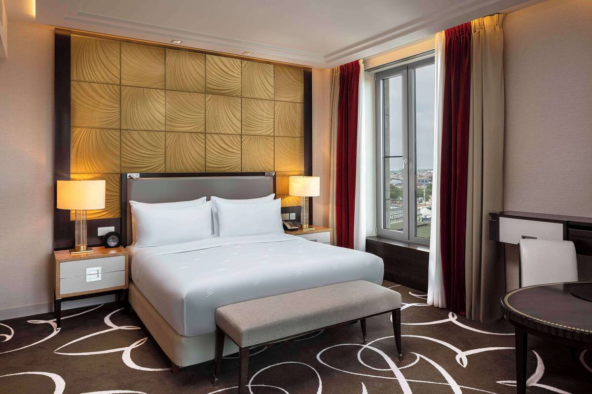Habitación de hotel con una cama grande, dos mesitas de noche con lámparas, un banco, una mesita pequeña y una ventana con cortinas.