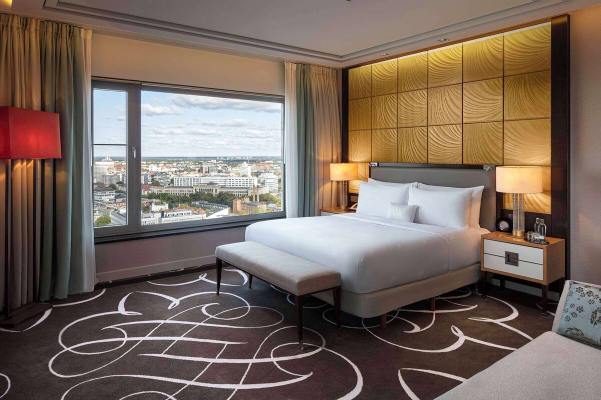 Una habitación de hotel con un ventanal grande, una cama, un banco y una mesita de noche con lámpara.