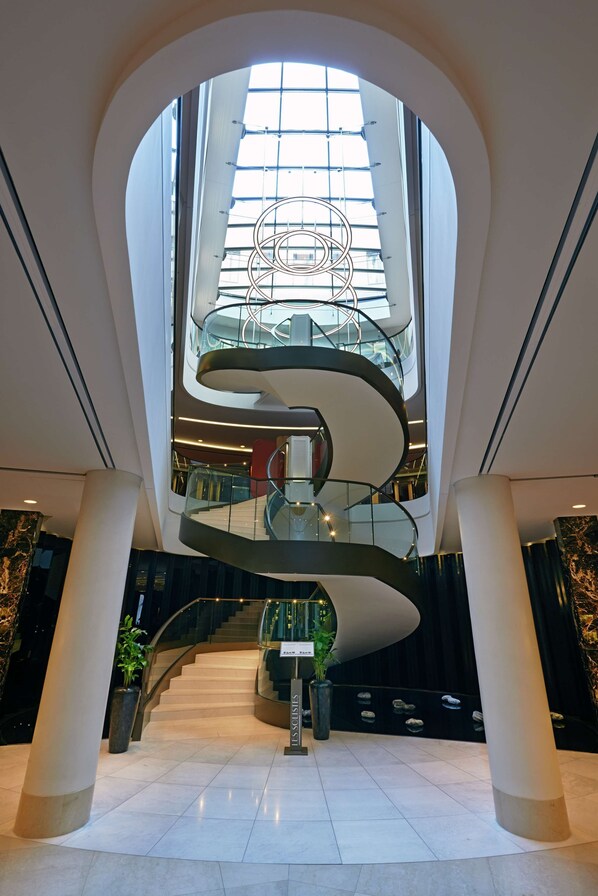 Un interior moderno con una escalera espiral central, barandillas de vidrio y un techo alto con tragaluces.