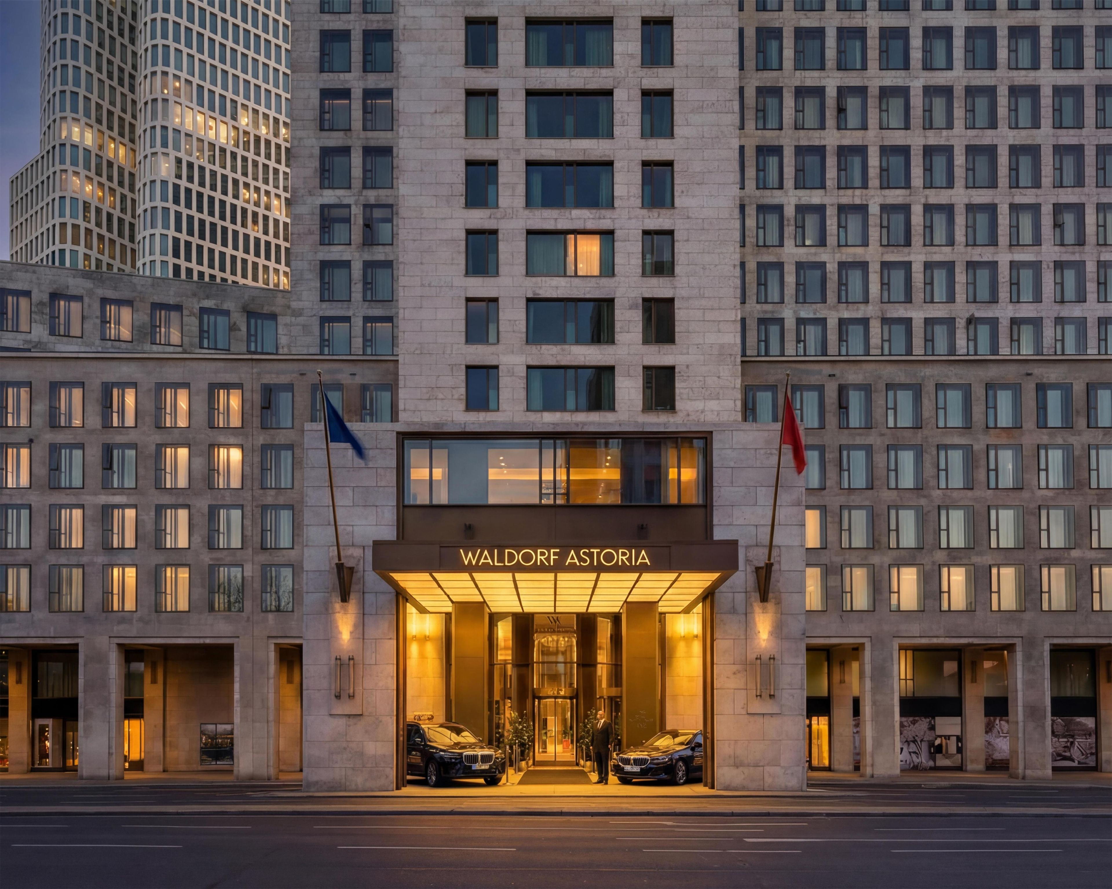 Een hotelgevel in moderne stijl, met twee geparkeerde auto's ervoor en vlaggen aan beide kanten.