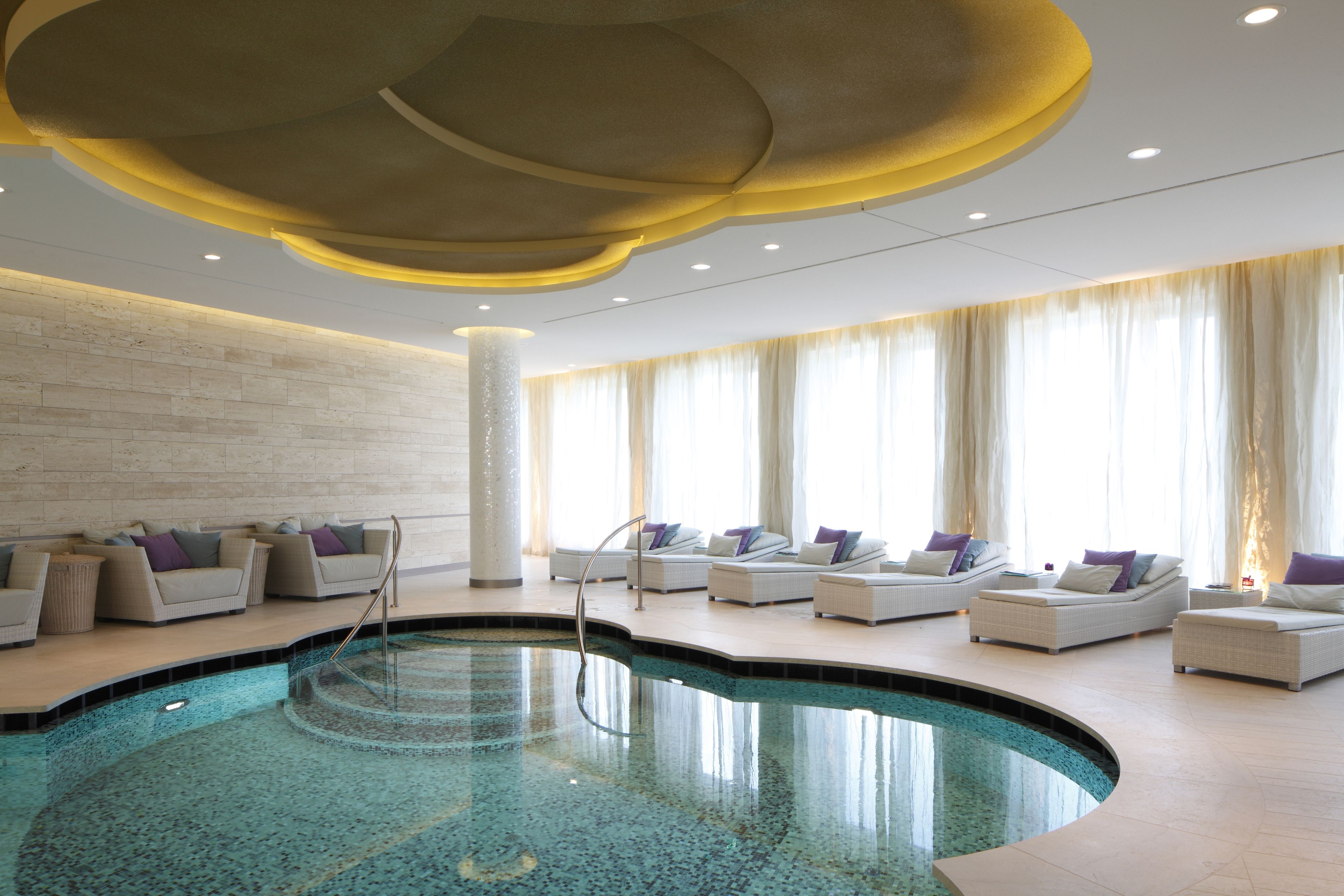 Un área de piscina interior moderna con sillones de descanso y un diseño de techo singular.