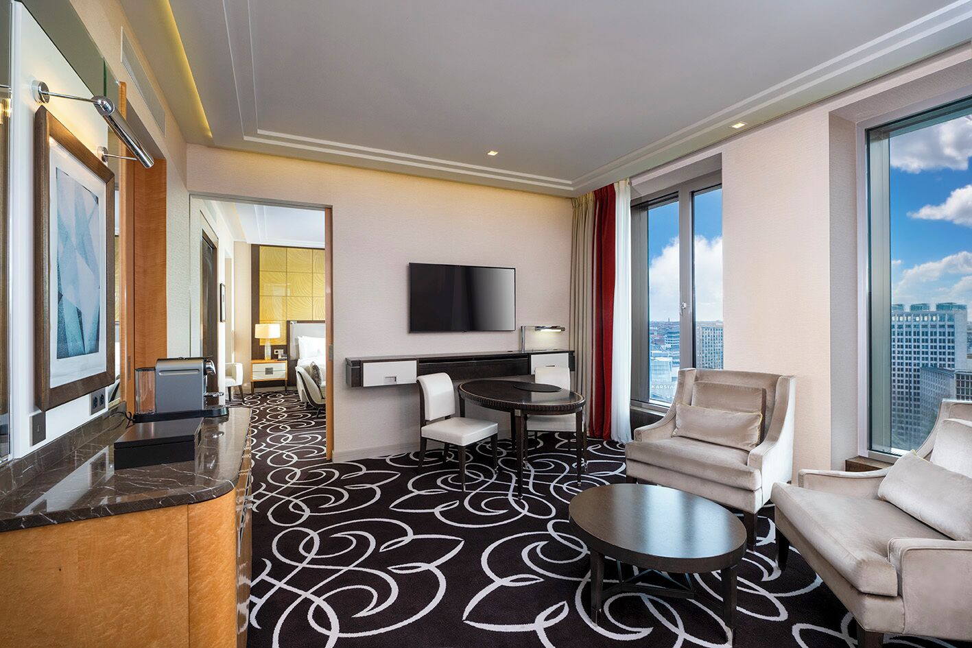 Habitación de hotel con un ventanal grande, un televisor de pantalla plana, una mesa redonda de comedor, dos sillones y un sofá.