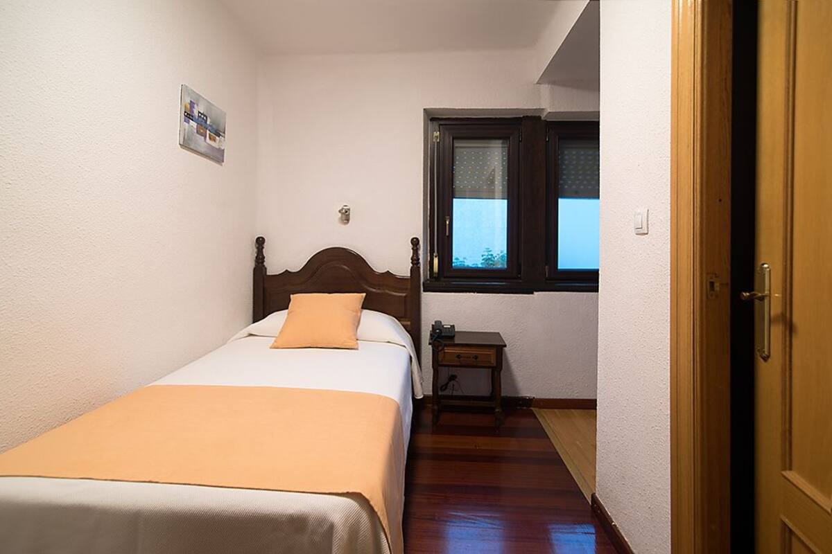 A bedroom with a single bed, a nightstand, a window with blinds, and a door.