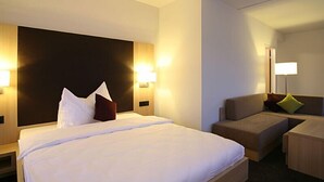 Hotelkamer met een groot bed, een bank en een bureau. Er zijn wandlampen en een nachtkastje met een lamp.