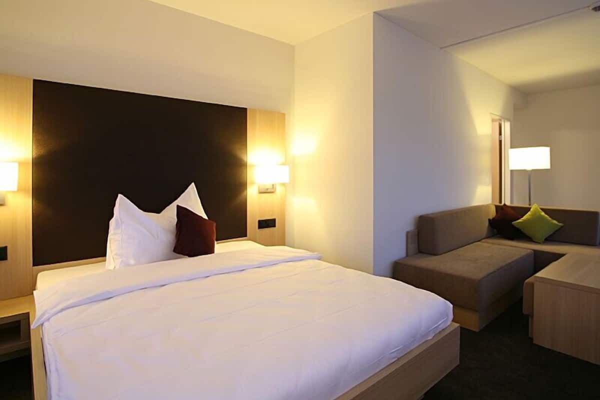 A hotel room with a large bed, a sofa, and a desk. There are wall-mounted lamps and a nightstand with a lamp.