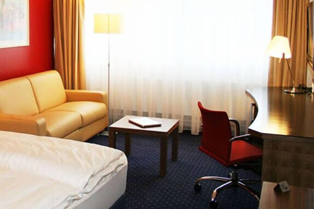 A hotel room with two beds, a sofa, a desk, and a chair. There are lamps on the desk and wall, and a window with curtains.