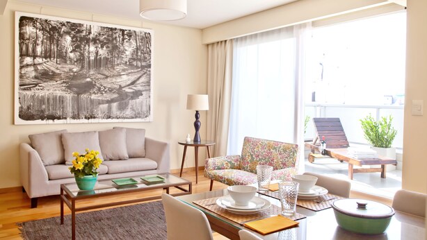 A modern living room with a sofa, coffee table, and a dining area. There is a large window providing natural light and a view of the outdoors.