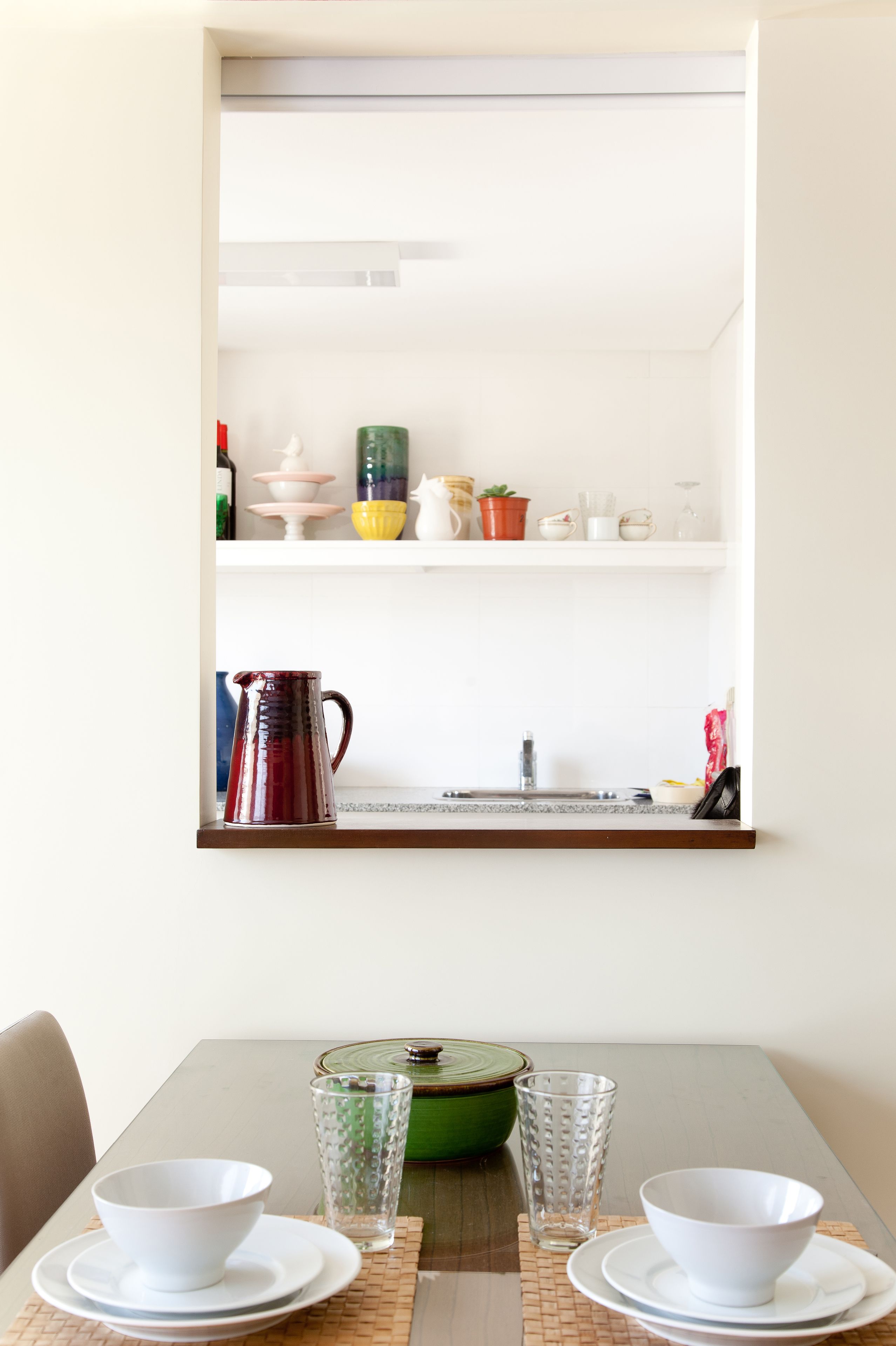 A dining area with a table set for two, a kitchen visible through a window, and a green bowl on the table.